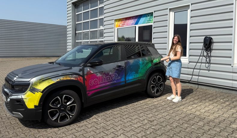 Ein SUV mit einer auffälligen, bunten Fahrzeugfolierung steht vor dem Gebäude der werbeatelier collinet GmbH und wird an einer Ladestation angeschlossen. Eine junge Frau steht lächelnd neben dem Fahrzeug.